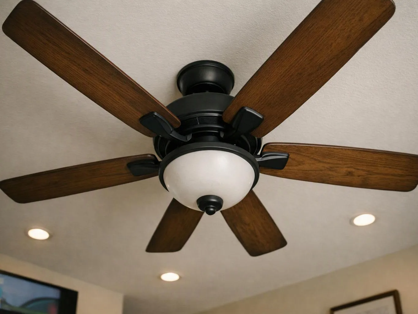 A ceiling fan with wood blades and a frosted light hangs in a room with recessed ceiling lights