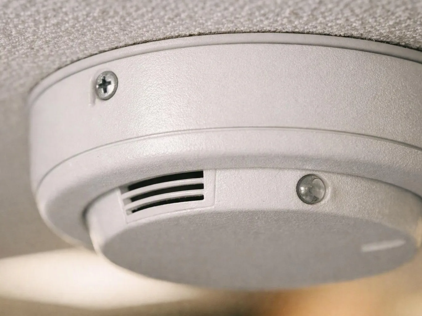 Close-up of a ceiling smoke detector with vents and screws mounted against a textured ceiling